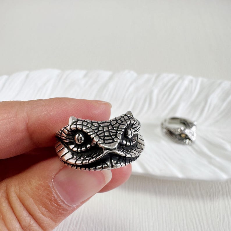 May include: A silver ring with an owl head design. The ring has textured detailing and large, round eyes. Another ring with the same design is visible in the background, resting on a white surface.