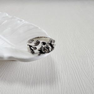 May include: A silver-toned ring featuring a detailed cat design. The ring wraps around the finger with the cat's head and paws forming the focal point. The ring is displayed on a white, feather-shaped dish.