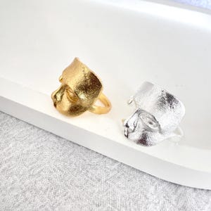 May include: Two rings, one gold and one silver, each with a sculpted face design. They are displayed on a white tray against a textured grey background. The face design is detailed and artistic, showcasing a unique style.