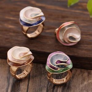 Puede incluir: Cuatro anillos dorados con diseños florales coloridos y superpuestos. Cada anillo presenta una combinación de colores diferente, incluyendo rosa, blanco, azul y morado. Los anillos están sobre una superficie de madera oscura.