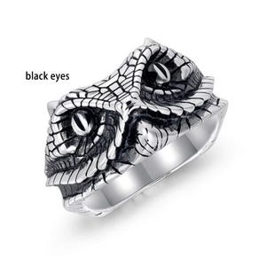 May include: A silver ring with a detailed snake head design. The snake's head is textured with scales and has black eyes. The ring has a polished finish and a wide band.