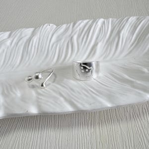 May include: Two silver rings on a white, textured surface. One ring features a curved design with two small spheres. The other is a wider band with a cut-out design. The rings are likely made of sterling silver.
