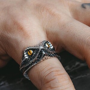May include: A silver snake head ring with detailed scales and two yellow eyes. The ring is worn on a finger, highlighting the intricate design. The jewellery piece is crafted with attention to detail.