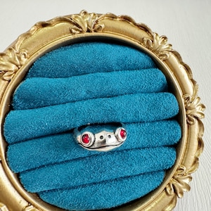 May include: A silver frog ring with red gemstone eyes sits on a teal velvet display. The ring is set in a gold-coloured, ornate, oval-shaped frame. The frog's face is detailed with two round red gems for eyes and a simple mouth.