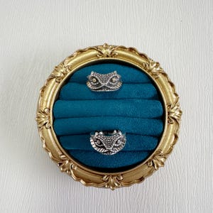 May include: Two silver rings, each with a snake head design, are presented on a teal velvet ring holder within a gold ornate frame. The rings feature detailed scale and eye patterns. The frame has a vintage style.