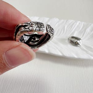 May include: A silver snake ring with intricate scale detailing and a hinged design. The ring is open, showing the inner details, and another snake head ring is in the background on a white surface.