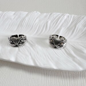 May include: Two silver rings featuring a snake head design. The rings have detailed scale patterns and small, gold-coloured accents for the eyes. They are displayed on a white, textured surface.