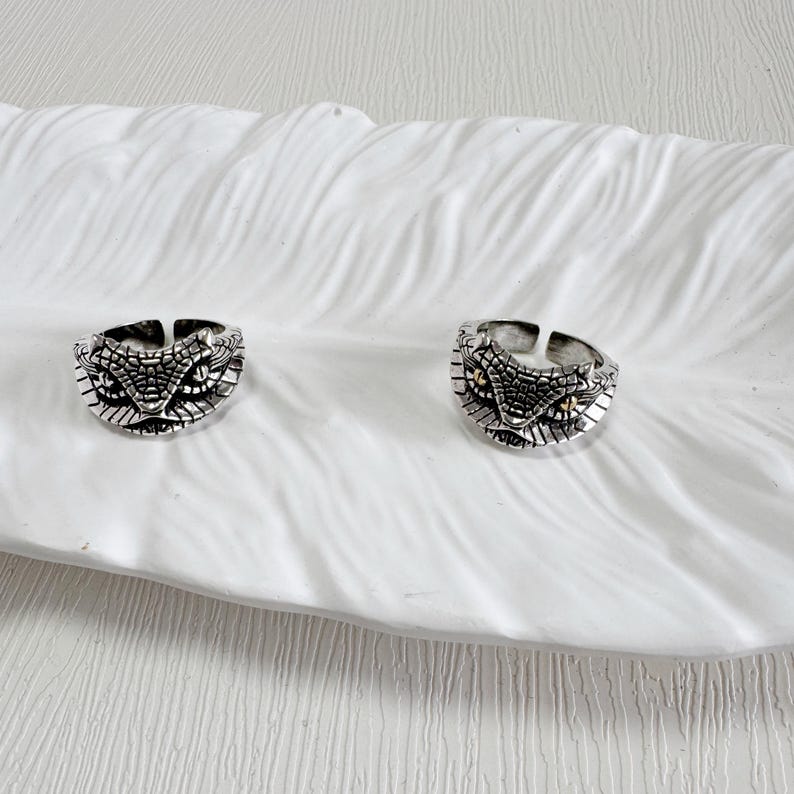 May include: Two silver rings featuring a snake head design. The rings have detailed scale patterns and small, gold-coloured accents for the eyes. They are displayed on a white, textured surface.