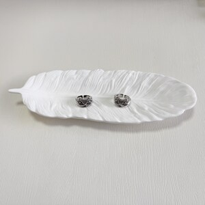 May include: Two silver rings with intricate designs sit on a white, feather-shaped ceramic dish. The rings have a dark, oxidised finish, and the dish has a textured surface. The rings are likely adjustable.