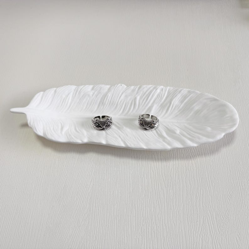 May include: Two silver rings with intricate designs sit on a white, feather-shaped ceramic dish. The rings have a dark, oxidised finish, and the dish has a textured surface. The rings are likely adjustable.