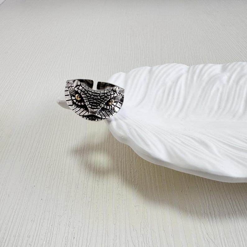 May include: A silver ring with a snake head design. The ring features detailed scales and small, gold-coloured eyes. The ring is displayed on a white, feather-shaped dish.