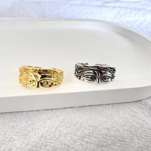 Puede incluir: Dos anillos, uno dorado y otro plateado, cada uno con un diseño de rostro esculpido. Los anillos se exhiben en una bandeja blanca sobre un fondo neutro. El diseño del rostro incluye ojos, nariz y cabello.