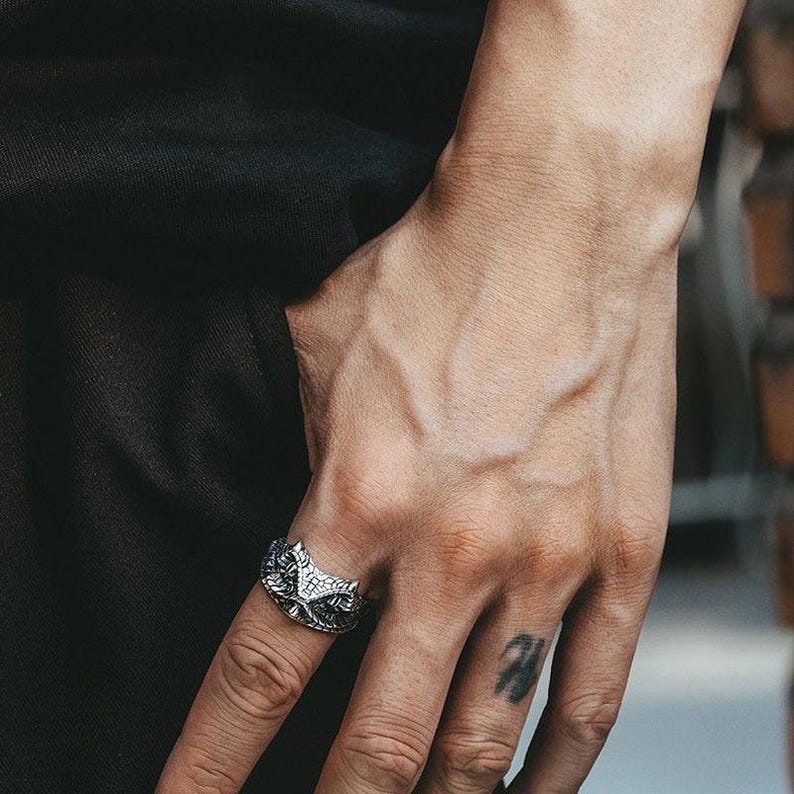 May include: A silver ring with an owl design. The ring has detailed features, with the owl's face and feathers clearly visible. The ring is worn on a finger, showcasing the craftsmanship. The ring is a piece of jewellery.