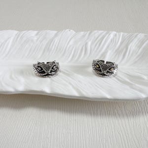 May include: Two silver snake head rings with intricate scale detailing and gold-coloured eye accents are shown on a white, feather-shaped dish. The rings are adjustable, with a vintage or gothic style.