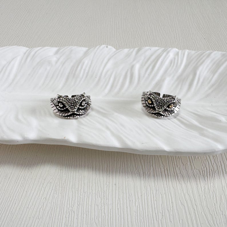 May include: Two silver snake head rings with intricate scale detailing and gold-coloured eye accents are shown on a white, feather-shaped dish. The rings are adjustable, with a vintage or gothic style.