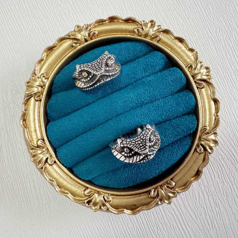 May include: Two silver rings, each with a snake head design, rest on a teal velvet ring holder within a gold ornate frame. The rings feature detailed scales and eye features.