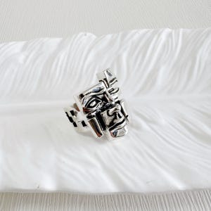 May include: A silver-toned ring with a geometric face design. The ring features a stylized face with defined features, including eyes, a nose, and a mouth. The ring is set against a white, textured background.