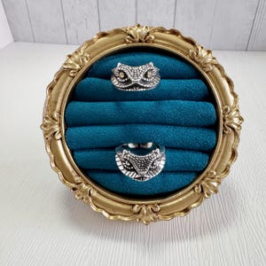 May include: Two silver snake head rings displayed on a teal velvet ring holder within a gold ornate frame. The rings have detailed scale and eye designs. The frame has a decorative, vintage style, reminiscent of a picture frame.