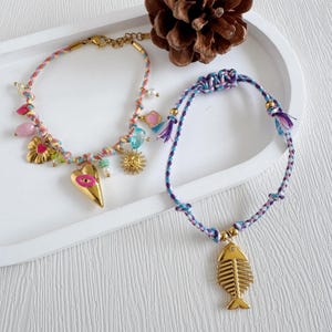 May include: Two colourful handmade bracelets. One features a gold heart charm with an eye, a sun, and other charms. The other has a gold fishbone pendant. Both are on a white tray with a pinecone.