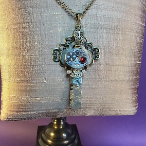 Può includere: Una collana con un ciondolo a forma di chiave d'argento, decorata con un quadrante dell'orologio e accenti di pietre preziose rosse. La collana è su una catena marrone ed è esposta su un manichino.