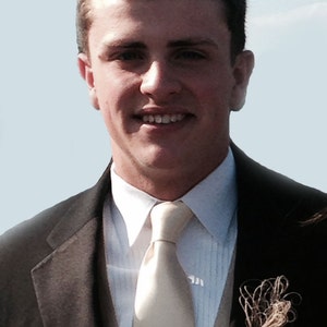Pu&ograve; includere: Un uomo con una giacca di abito marrone, una camicia bianca e una cravatta beige chiaro. Indossa un boutonni&egrave;re con una piuma marrone e uno stelo marrone.