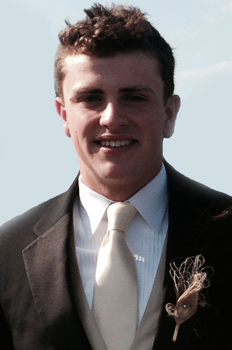 Pu&ograve; includere: Un uomo con una giacca di abito marrone, una camicia bianca e una cravatta beige chiaro. Indossa un boutonni&egrave;re con una piuma marrone e uno stelo marrone.