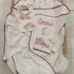 Può includere: Un set composto da un asciugamano con cappuccio, una bavaglino e un guanto da bagno bianchi, decorati con un motivo di nuvole e lune rosa e il nome Emma ricamato. Ogni pezzo presenta un bordo rosa metallizzato e una consistenza morbida.