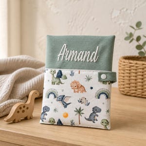 Puede incluir: Un estuche de tela rectangular con una sección superior en verde salvia que tiene el nombre Armand bordado en blanco. La parte inferior muestra un fondo blanco con un estampado de dinosaurios, arcoíris, soles y palmeras de colores.