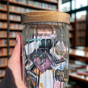 Könnte beinhalten: Ein klares Glasgefäß mit Holzdeckel, gefüllt mit gefalteten Papierzetteln. Das Gefäß ist gerippt und wird vor einem verschwommenen Hintergrund von Bücherregalen gehalten. Die Notizen scheinen Text und Bilder zu haben, was auf eine Sammlung von Erinnerungen hindeutet.