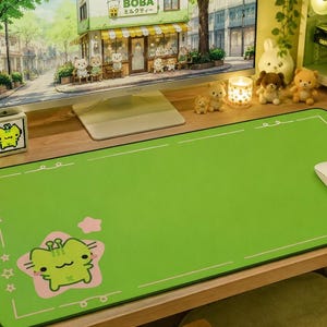 Puede incluir: Alfombrilla de ratón rectangular verde con borde blanco y diseño de rana de dibujos animados. La alfombrilla está sobre un escritorio de madera con un monitor de ordenador que muestra una escena de tienda Boba. Un ratón blanco está a la derecha.