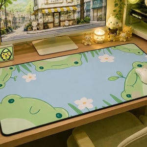 Op de afbeelding: Een bureau-opstelling met een computermonitor met een Boba-winkelscène. Een lichtblauwe muismat met groene kikkerillustraties en witte bloemen ligt voor de monitor. Een witte muis ligt op de muismat. Diverse decoratieve items omringen de monitor.