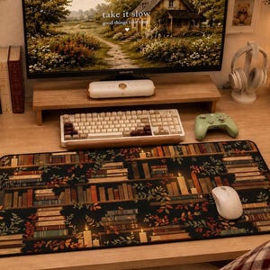 Op de afbeelding: Een rechthoekige bureaumat met een donker, herhalend patroon van boekenplanken gevuld met diverse boeken, brandende kaarsen en verspreide herfstbladeren. De mat ligt op een houten bureauoppervlak als ondergrond voor een computermuis.