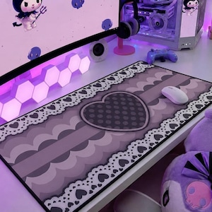 Puede incluir: Una alfombrilla de ratón rectangular morada y gris con un diseño de corazón y un ratón blanco. La alfombrilla tiene un borde de encaje con recortes de corazón. El fondo incluye un monitor de ordenador, una torre de ordenador y otros accesorios de juego.