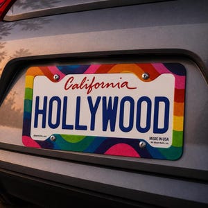 Puede incluir: Marco de matrícula con diseño de arcoíris y la palabra "HOLLYWOOD" en letras azules. La palabra "California" está en escritura roja. Fijado a un coche.