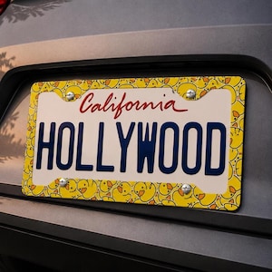 Puede incluir: Una placa de matrícula de California con la palabra "HOLLYWOOD" en letras mayúsculas azul marino. La palabra "California" está escrita en cursiva roja arriba. La placa tiene un borde amarillo con un patrón de patitos amarillos.