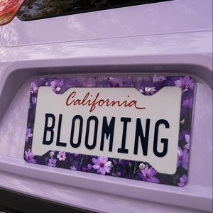 Puede incluir: Un marco de matrícula con un diseño floral con flores moradas y hojas verdes. La matrícula dice "BLOOMING" con "California" en escritura roja arriba. Montado en un coche lila.