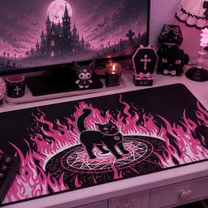 Puede incluir: Alfombrilla de escritorio negra con un gato negro dentro de un pentagrama rosa y llamas rosas. El fondo incluye un monitor de computadora con un castillo gótico, y objetos decorativos como una caja con forma de ataúd y una figura de gato negro.