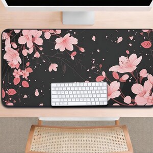 以下が含まれることがあります： ピンクの桜の花と葉のフローラルデザインが施された黒いデスクマット。白いキーボードがマットの上に置かれ、背景にはコンピューターモニターとマウスが見えます。長方形のマットは机の大部分を覆っています。