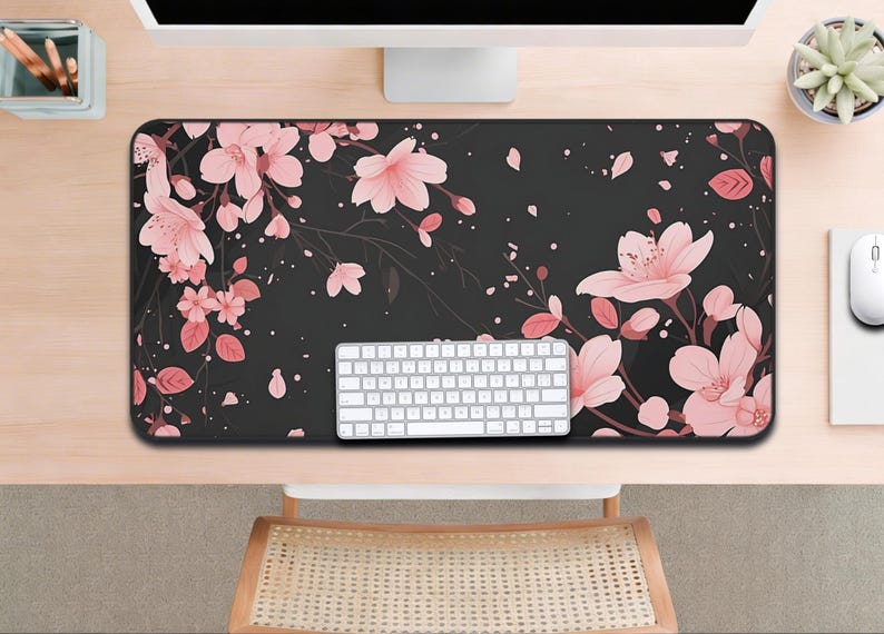 以下が含まれることがあります： ピンクの桜の花と葉のフローラルデザインが施された黒いデスクマット。白いキーボードがマットの上に置かれ、背景にはコンピューターモニターとマウスが見えます。長方形のマットは机の大部分を覆っています。