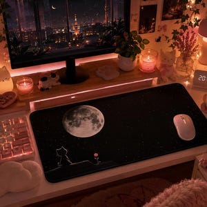 May include: A rectangular black desk mat featuring a large white full moon and a small silhouette of a cat sitting on a ledge looking at a single flower. The mat has a starry night sky pattern and is positioned on a desk with a computer mouse.