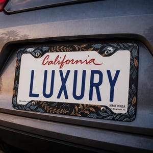 Puede incluir: Un marco rectangular para matrícula con fondo oscuro y un patrón repetitivo de hojas de helecho azules y doradas. El marco rodea una matrícula blanca de California con la palabra LUXURY escrita en letras mayúsculas azules.