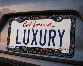 May include: A rectangular number plate surround featuring a dark background with a repeating pattern of blue and gold fern leaves. The frame surrounds a white California number plate with the word LUXURY in large blue capital letters.