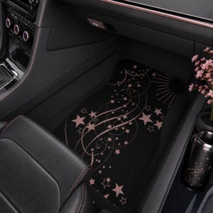 May include: A black car floor mat featuring a celestial pattern of light pink stars, swirling lines, and sunburst rays. The design is printed across the rectangular mat, which is positioned on the floor of a vehicle interior with dark upholstery.
