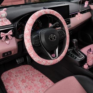 Op de afbeelding: Een stuurwielhoes voor in de auto met een roze kersenbloesempatroon, gecombineerd met bijpassende interieuraccessoires, waaronder een vloermat, een roze geruite strik, een klein roze hartvormig embleem, een hangende bloemhanger en een roze beker.
