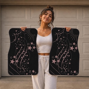 May include: A pair of rectangular black car floor mats featuring a celestial design with light pink stars, swirling lines, and sunburst rays. The mats have rounded corners and a durable texture, showcasing a consistent pattern of scattered stars.