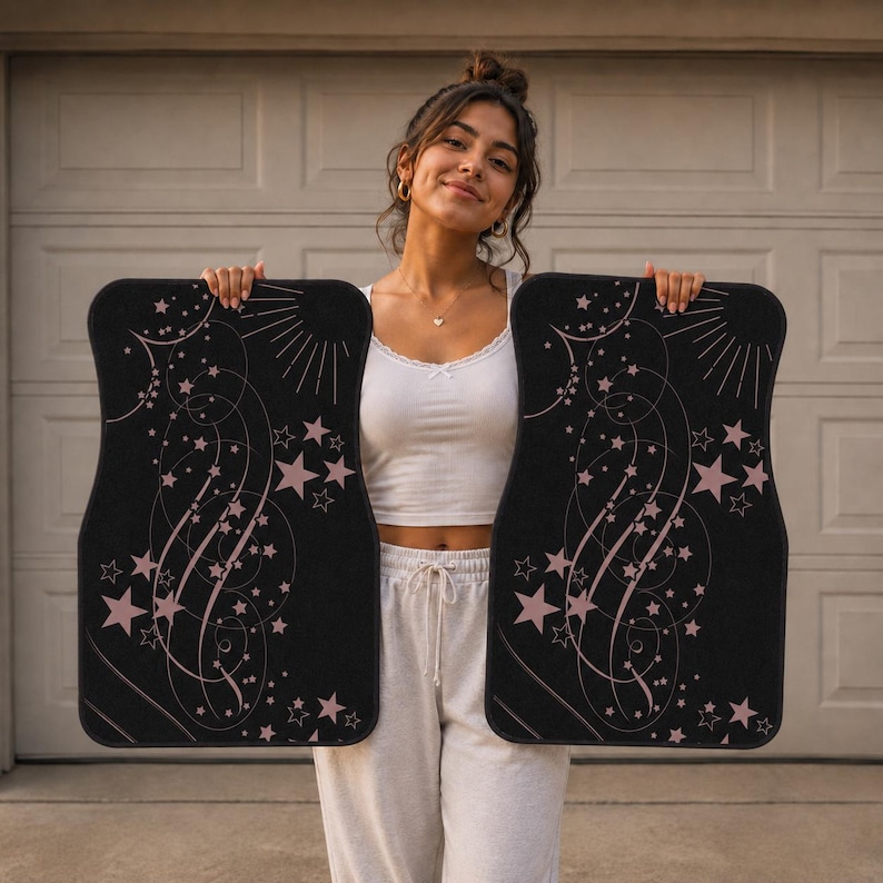 May include: A pair of rectangular black car floor mats featuring a celestial design with light pink stars, swirling lines, and sunburst rays. The mats have rounded corners and a durable texture, showcasing a consistent pattern of scattered stars.