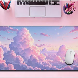 Puede incluir: Una alfombrilla de ratón rectangular con una escena de cielo de ensueño con nubes esponjosas, rosas y moradas contra un cielo azul. Un ratón de ordenador blanco descansa sobre la alfombrilla. La alfombrilla está sobre un escritorio rosa con un portátil y material de oficina.