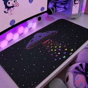 Puede incluir: Alfombrilla de ratón rectangular negra con un diseño de OVNI morado que emite corazones coloridos sobre un fondo estrellado. La alfombrilla está diseñada para uso informático y añade un toque decorativo a una configuración de escritorio. El diseño incluye un OVNI morado con detalles rojos y blancos.