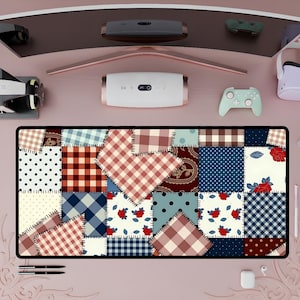 Puede incluir: Alfombrilla de escritorio rectangular con diseño de patchwork, que muestra cuadrados de varios patrones, incluyendo lunares, cuadros vichy y estampados florales en tonos rojos, azules y crema. La alfombrilla está rodeada de accesorios tecnológicos sobre un escritorio rosa.
