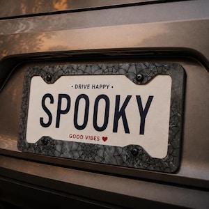 Puede incluir: Un marco de matrícula con un diseño de telaraña y la palabra "SPOOKY" en letras grandes y en negrita. El marco también incluye las frases "DRIVE HAPPY" y "GOOD VIBES" con un símbolo de corazón. El marco está sujeto a un vehículo.